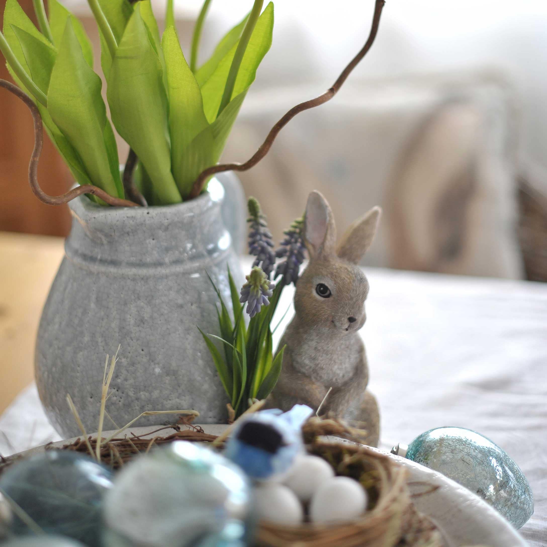 Grijze keramische vaas met groene bladeren en konijnenfiguur als paasdecoratie op tafel