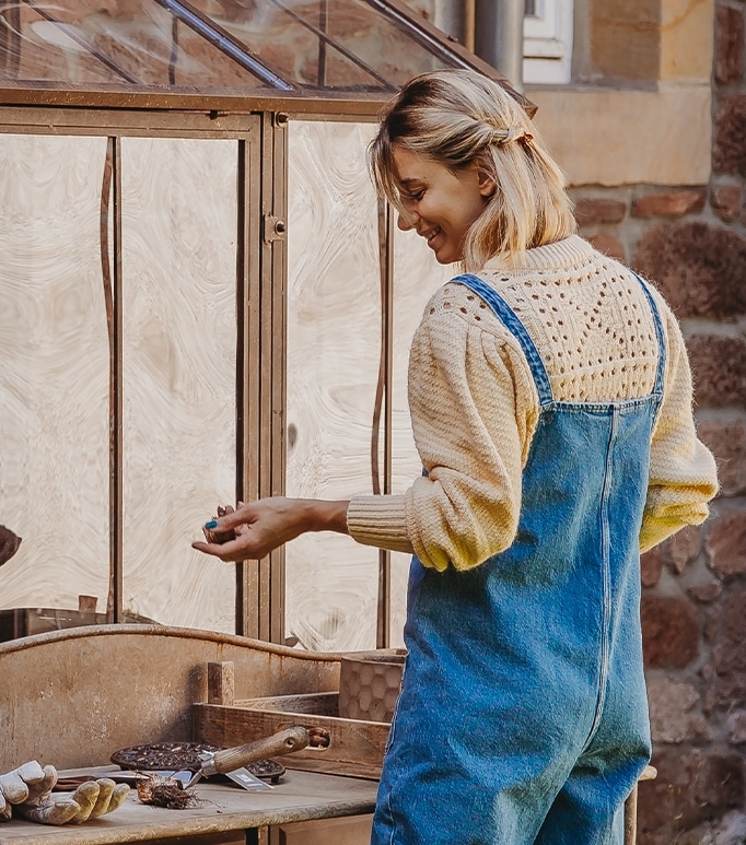 Vrouw in blauwe tuinbroek en crème trui werkt aan een houten tafel in de tuin