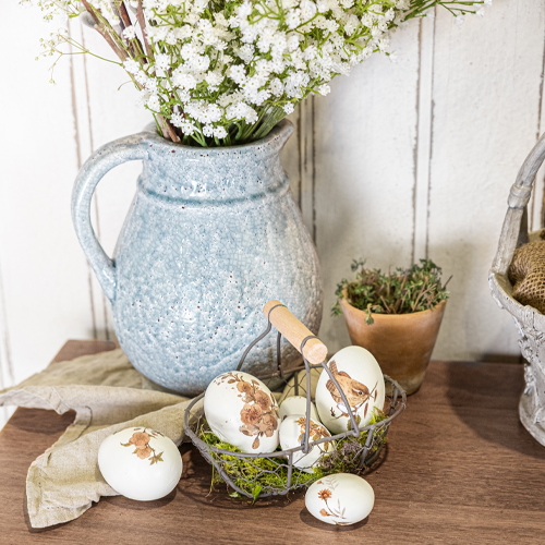 Blauwe keramische kan met witte bloemen en decoratieve eieren met dierenmotieven op houten tafel