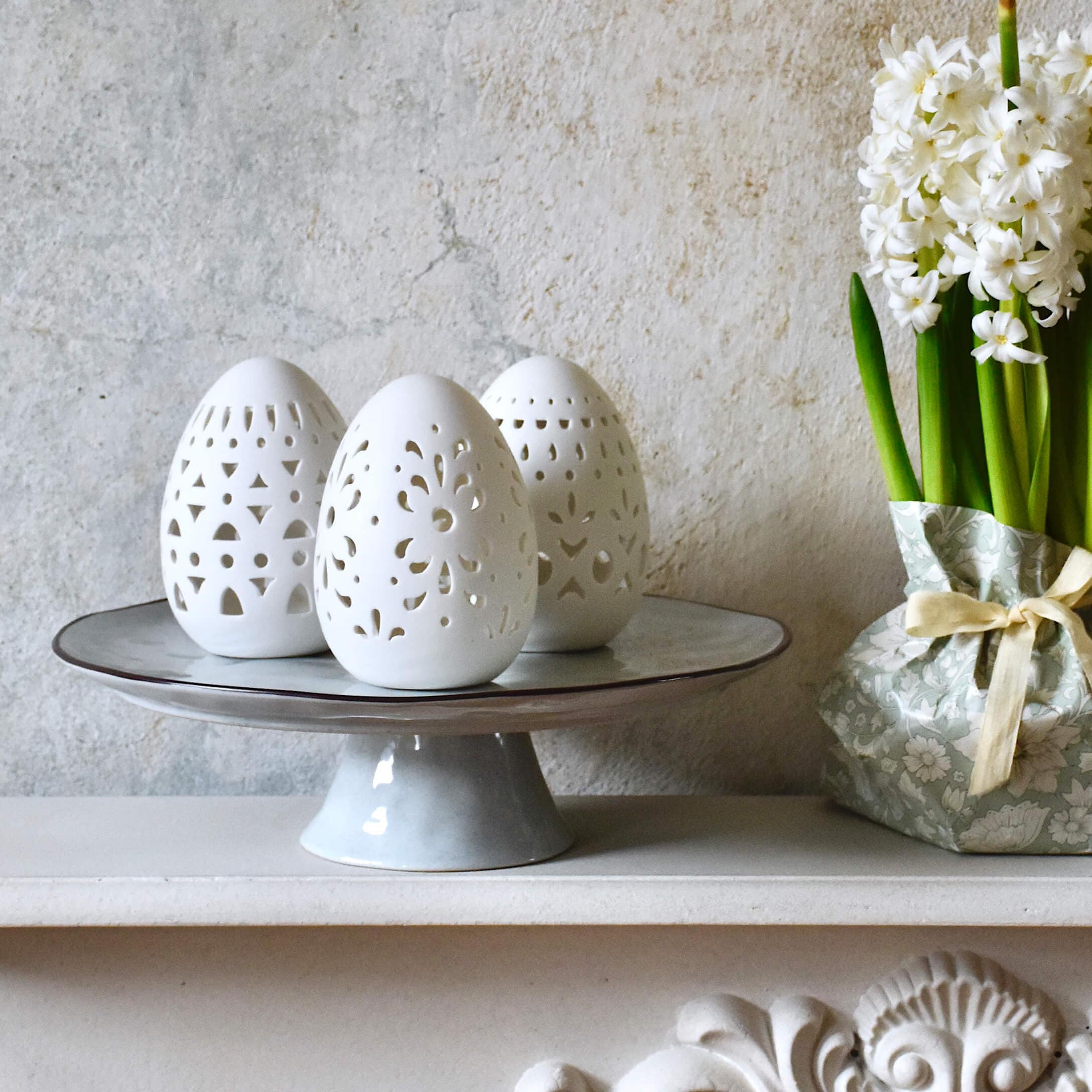 Witte keramische paaseieren met opengewerkt patroon op een grijze schaal naast witte bloemen in decoratieve verpakking