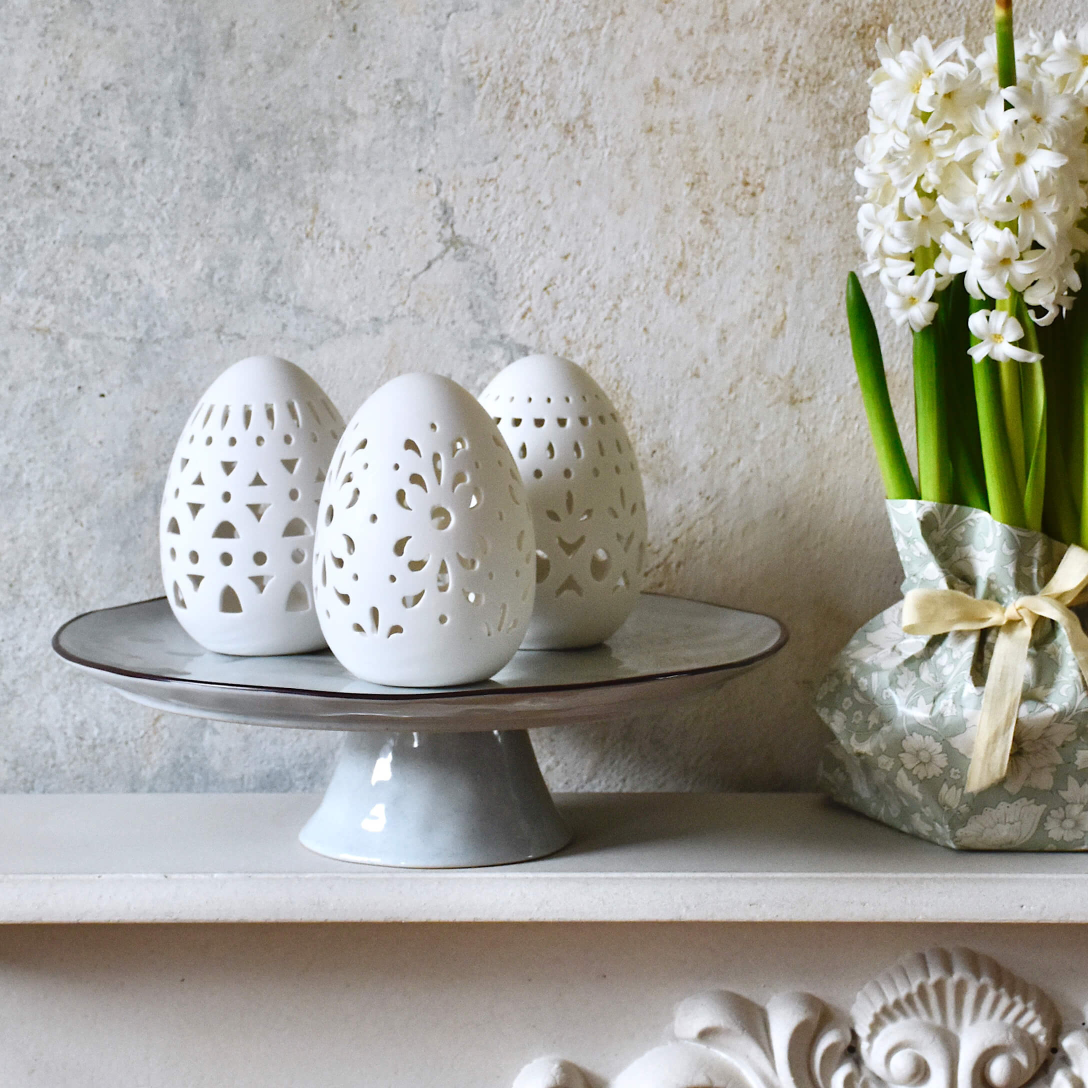 Witte keramische paaseieren met opengewerkt patroon op een grijze schaal naast witte bloemen in decoratieve verpakking