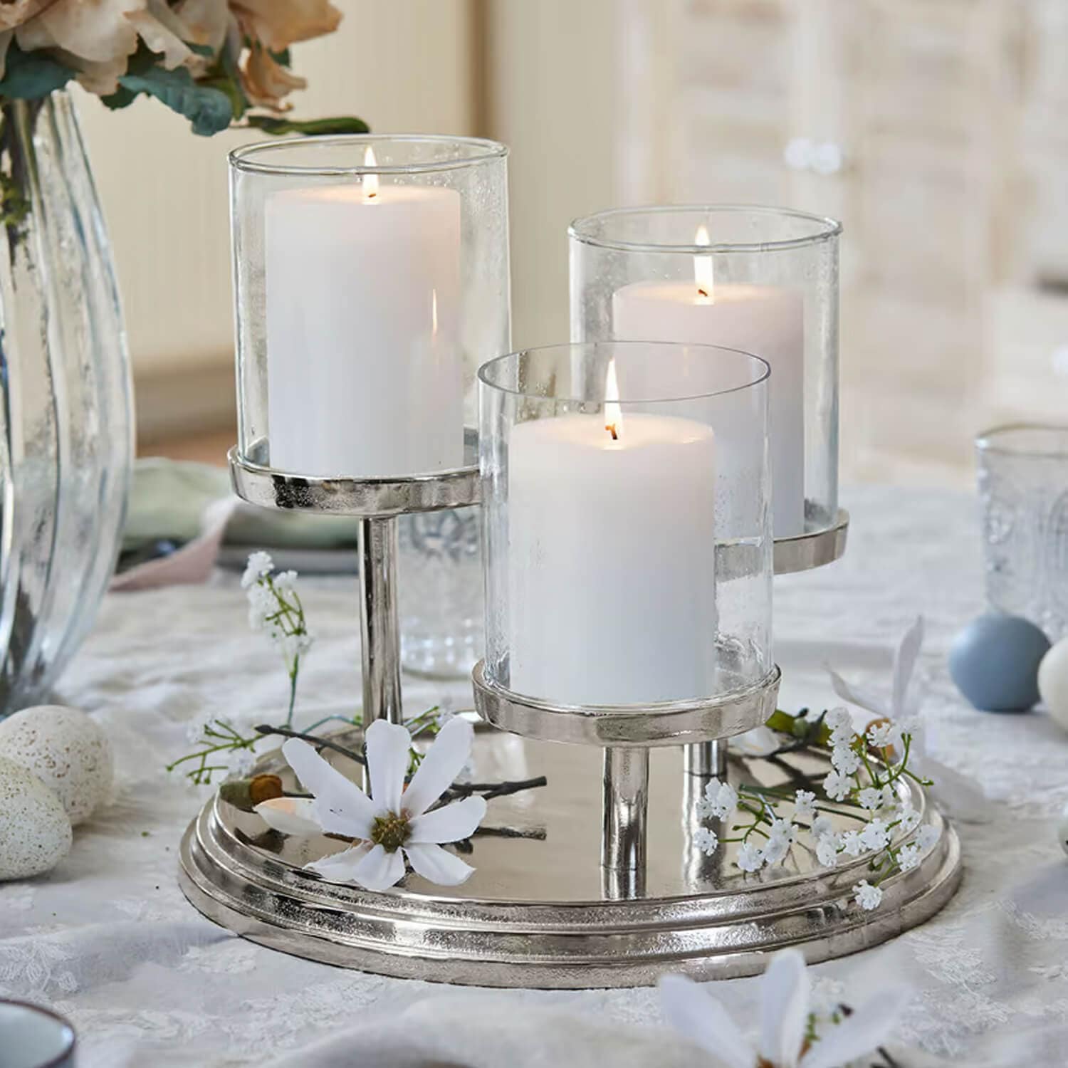 Driedelige kandelaar van zilverkleurig metaal met drie witte kaarsen en decoratieve bloemen op tafel