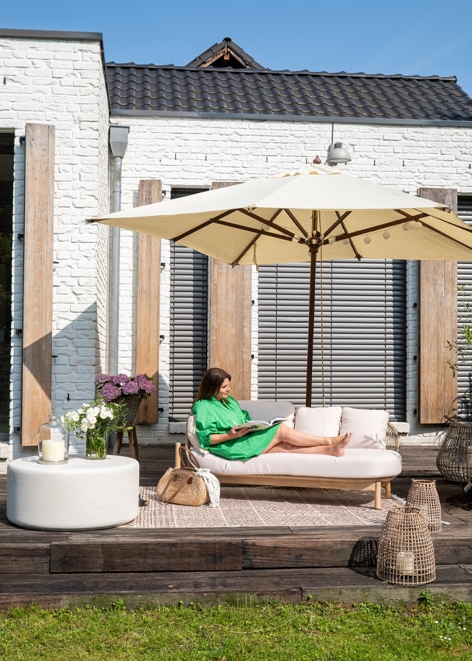 Vrouw in groene jurk ontspant op lichte bank onder crème parasol op houten terras