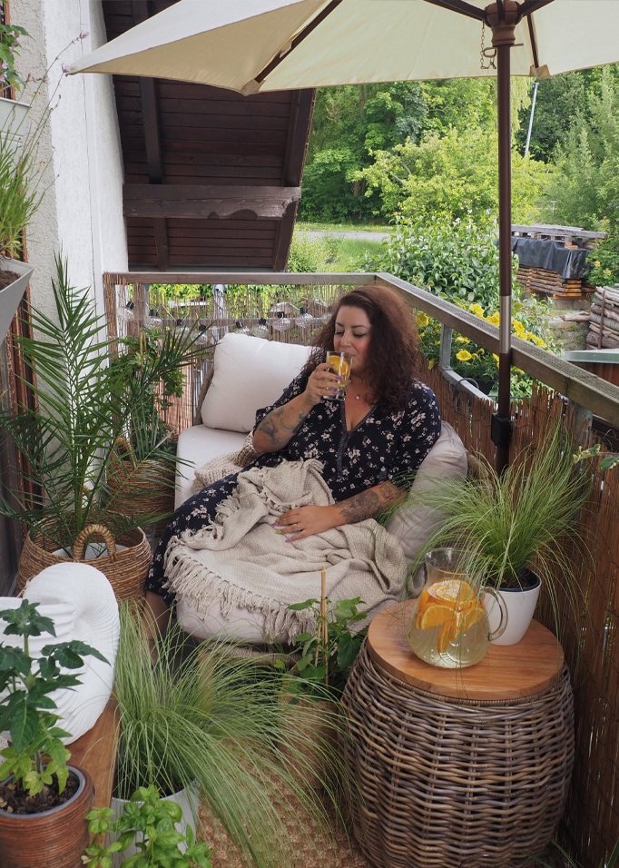 Vrouw ontspant op balkon met rotan tafel en crème deken, omringd door groene planten.