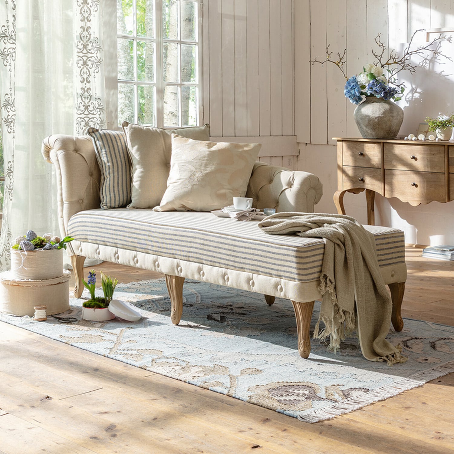 Beige gestoffeerde chaise longue met houten poten en lichte kussens in lichte woonkamer