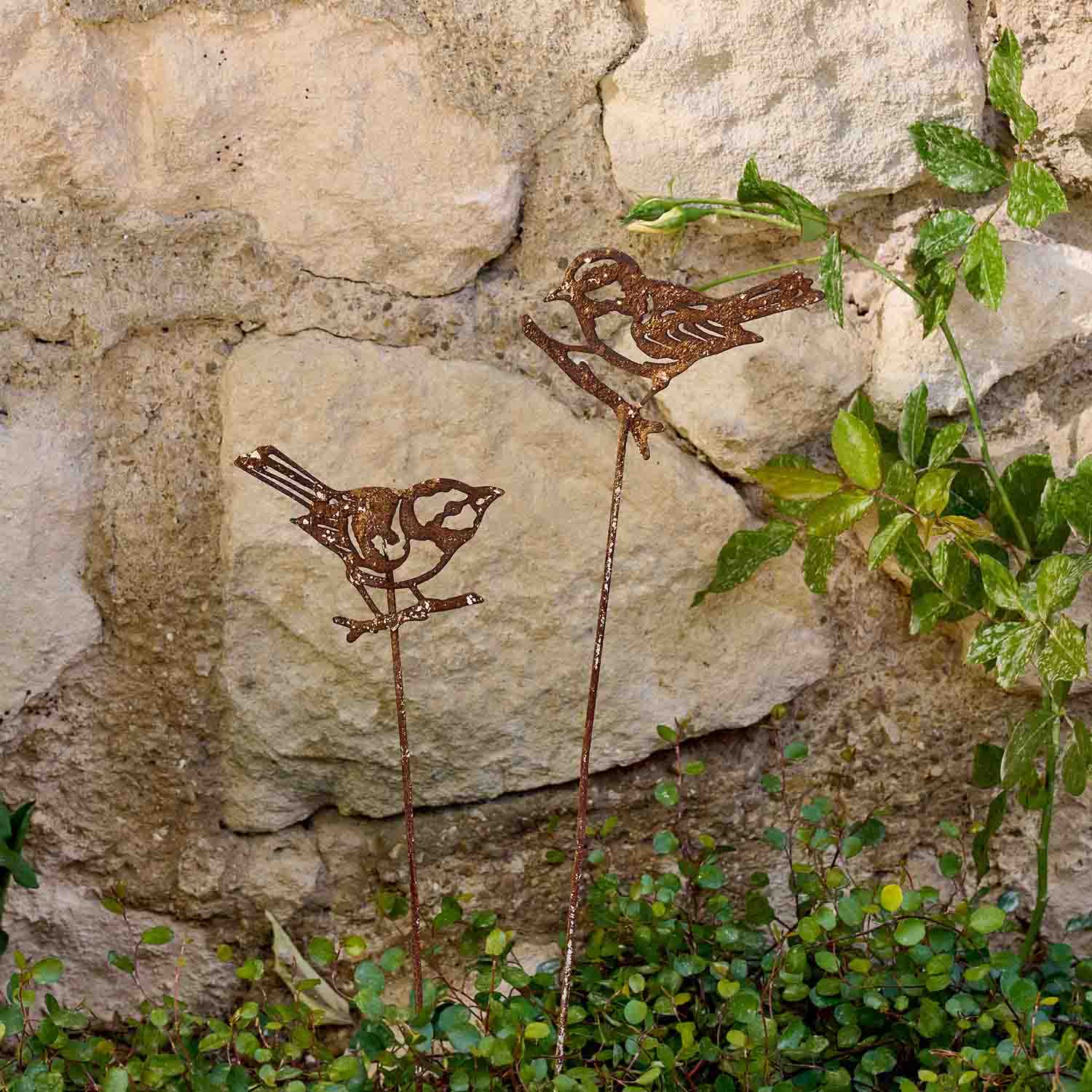 Decoratieve metalen vogels met roestkleurige afwerking op stokken in tuin bij stenen muur