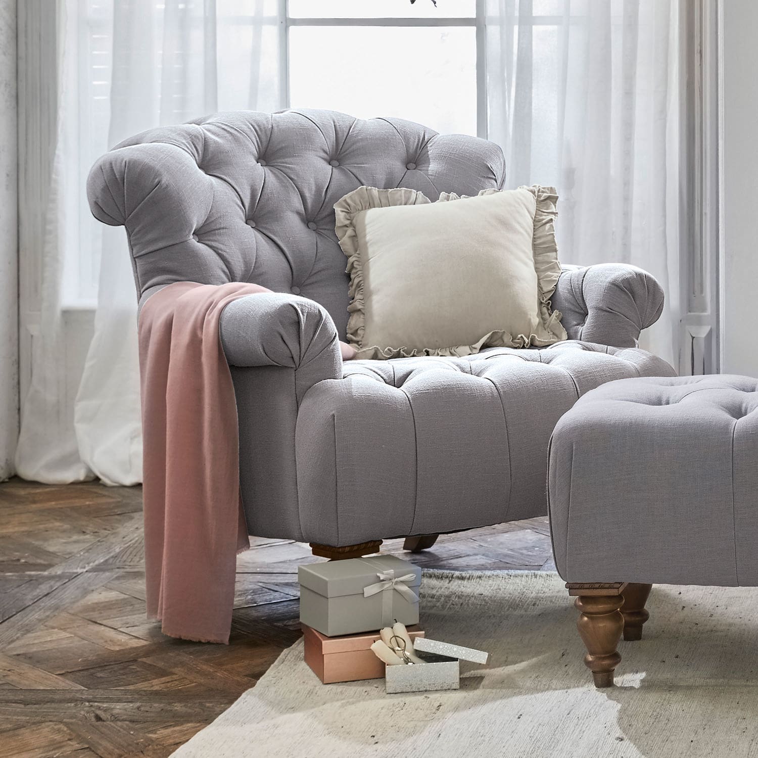 Fauteuil Springfield Grijze gestoffeerde fauteuil met voetenbank, crème kussen en roze deken op houten vloer in woonkamer