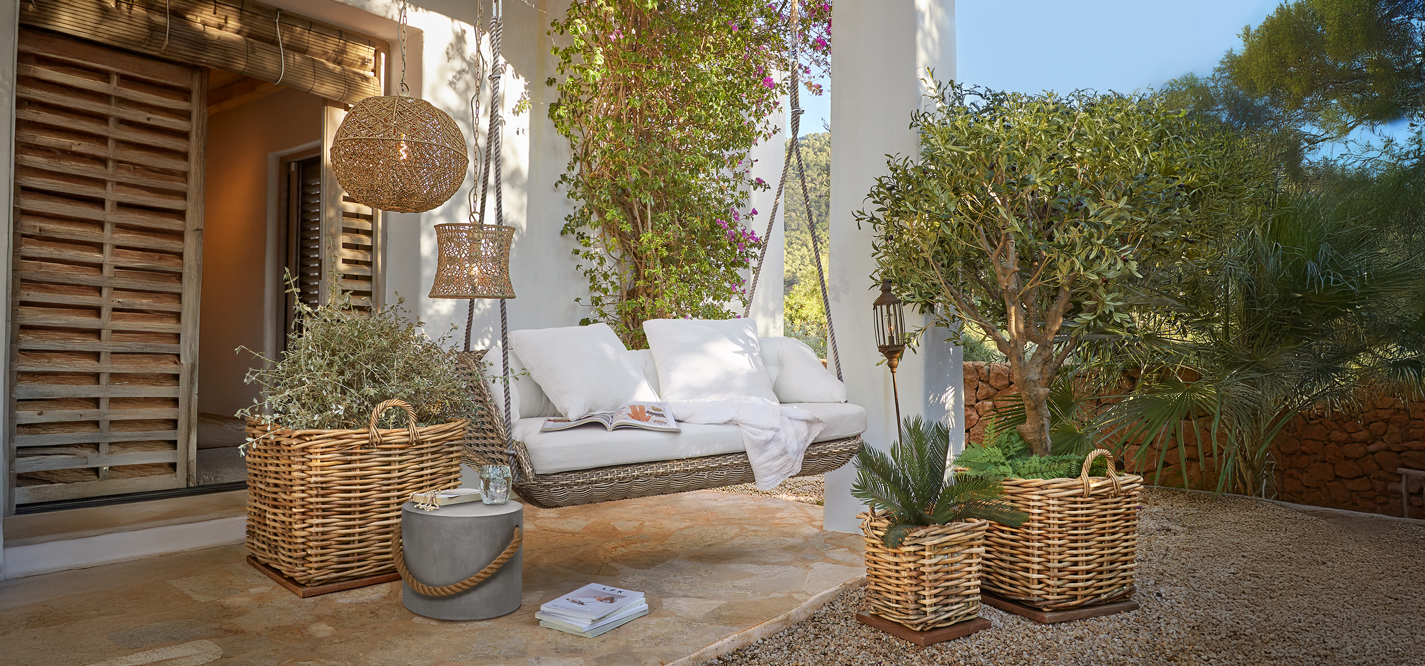 Rotan schommel met witte kussens op terras met rieten manden en groene planten