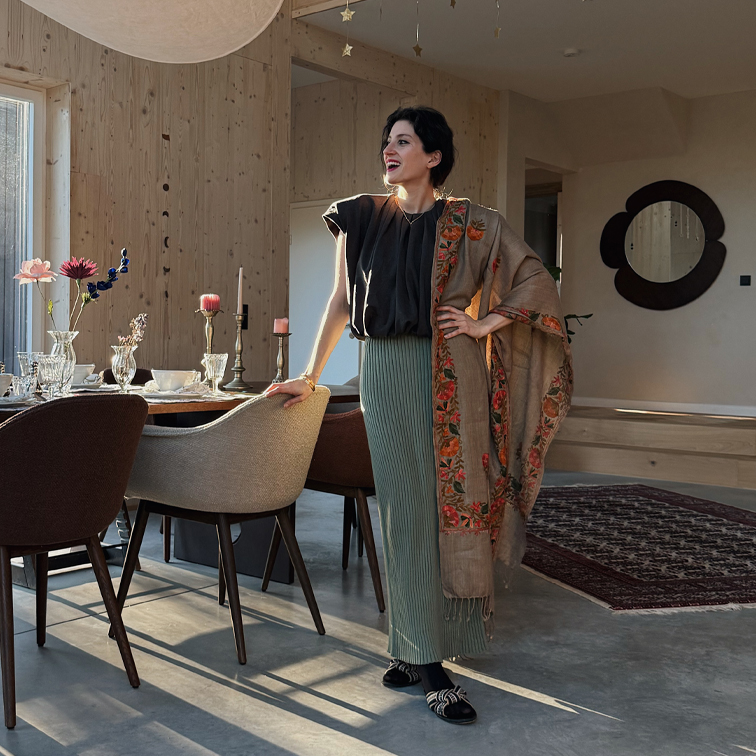 Vrouw in groene geplooide rok, zwarte top en beige sjaal met bloemenpatroon in modern interieur