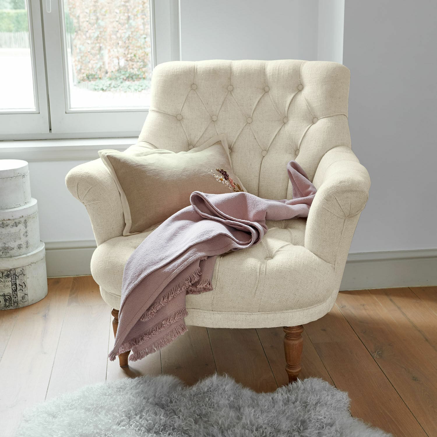 Fauteuil Rive Dorée Beige gestoffeerde fauteuil met houten poten, roze deken en grijs schapenvachtkleed op houten vloer