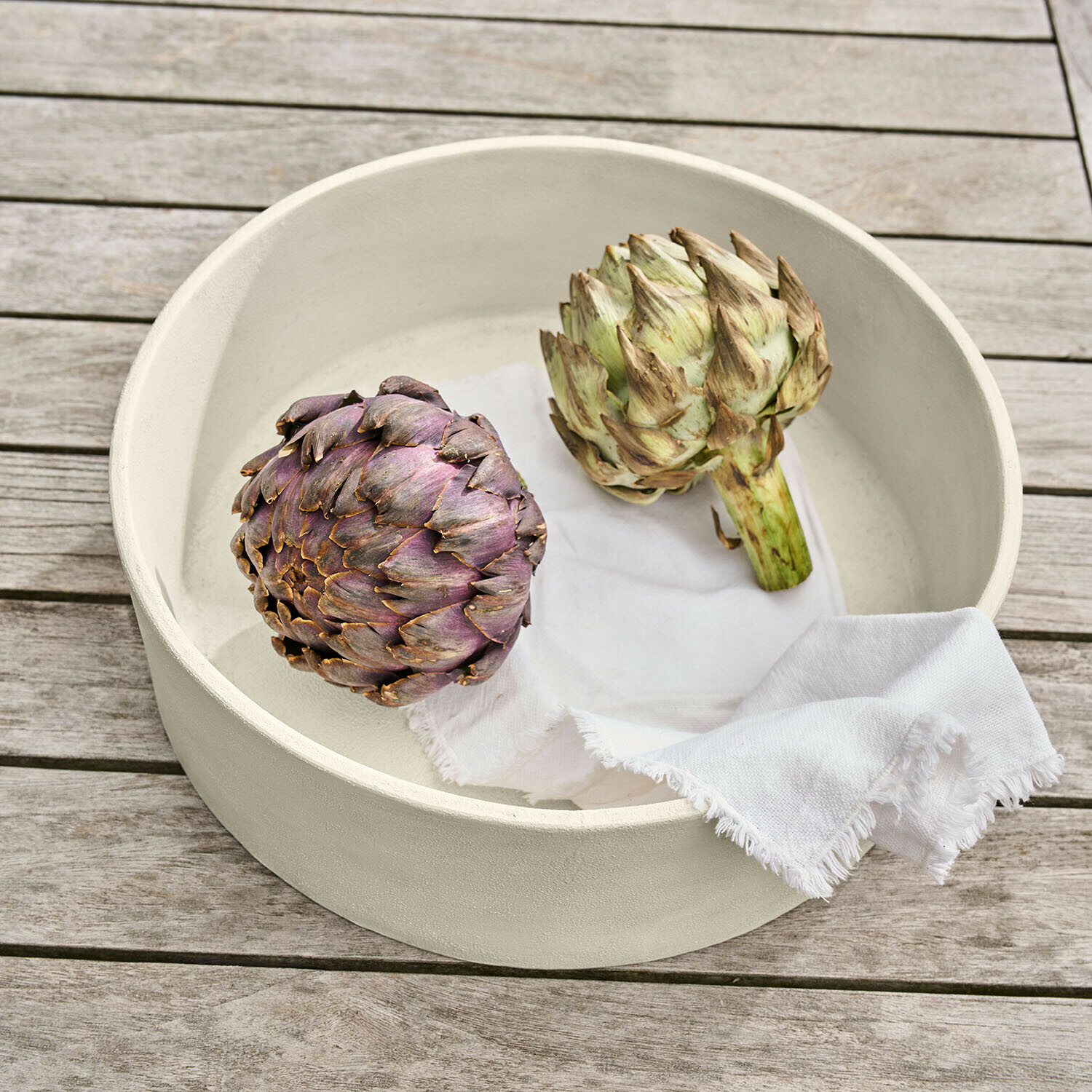 Ronde crème keramische schaal met twee artisjokken en een wit doek op een houten tafel
