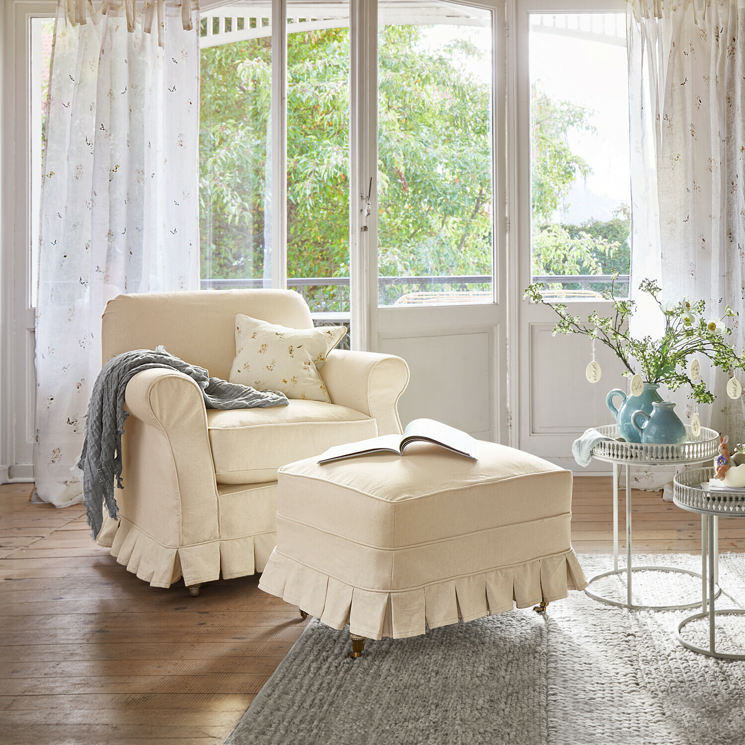 Beige stoffen fauteuil en voetenbank in lichte woonkamer met grote ramen en decoratieve tafel