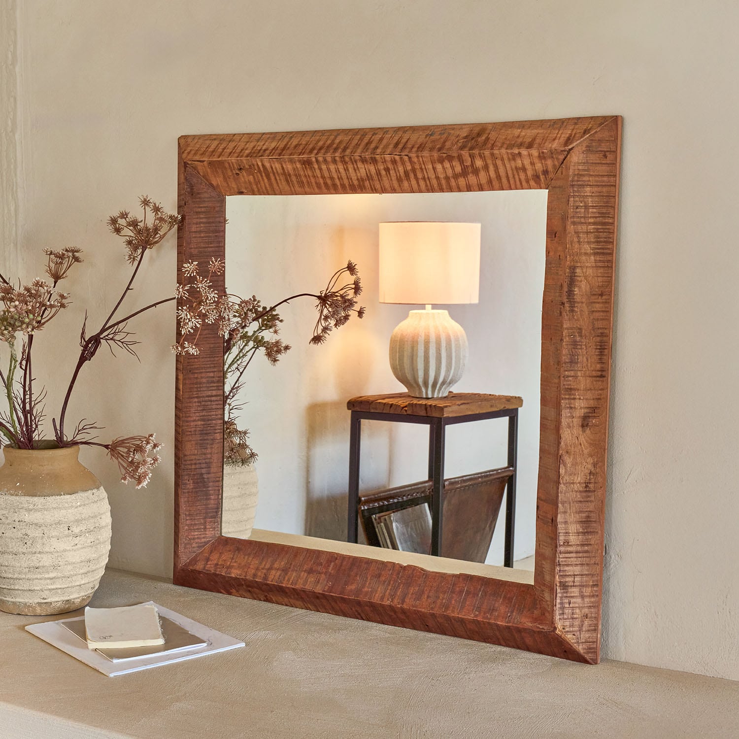Vierkante spiegel met donkere houten lijst aan de muur, reflecteert lamp en rustieke tafel