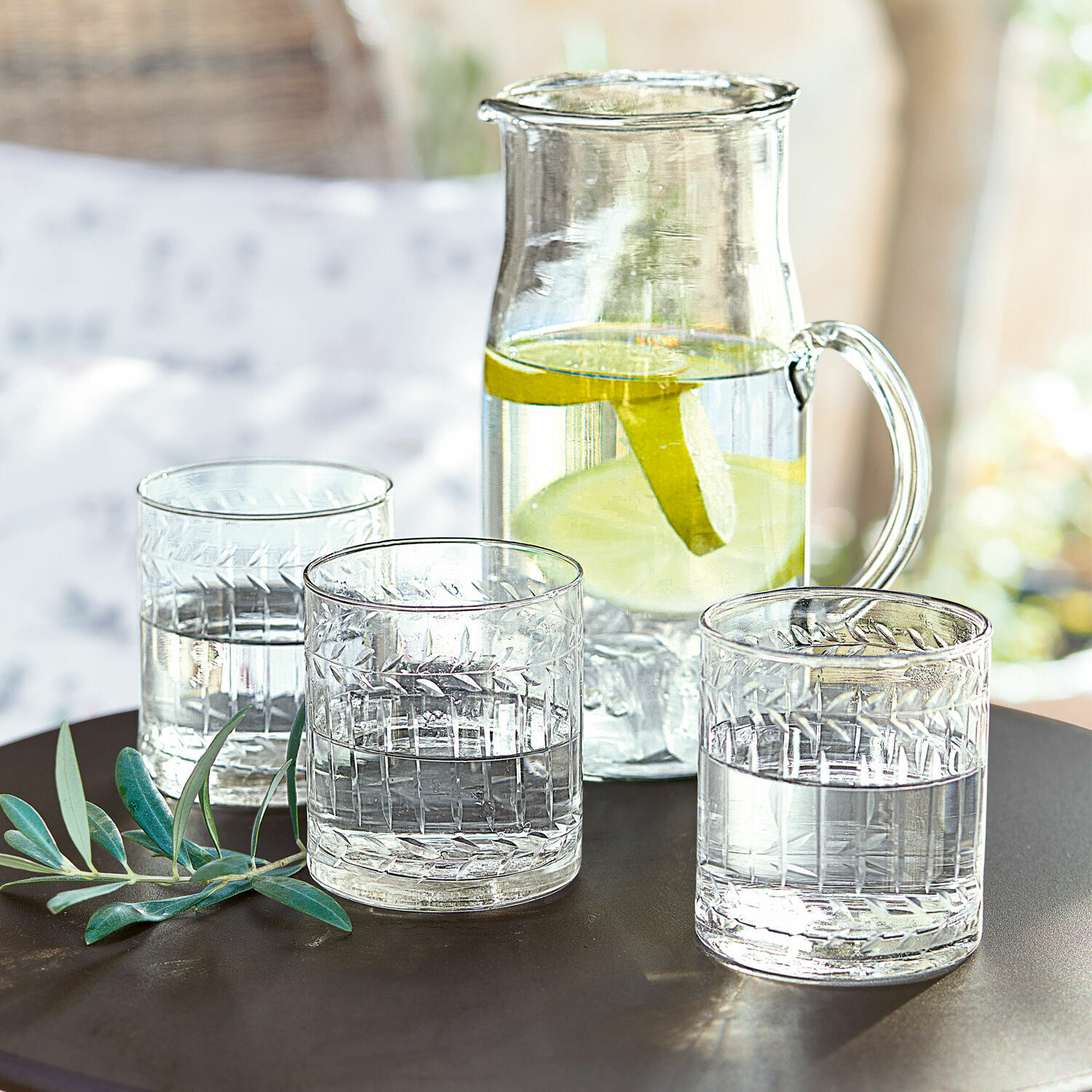 Glazen karaf met citroenplakjes en drie glazen water op een donkere tafel met groene bladeren als decoratie