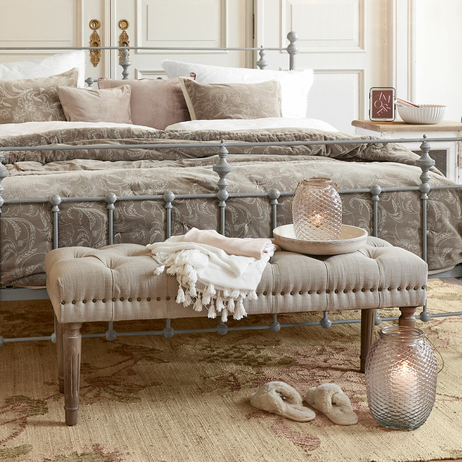 Grijs metalen bedframe met beige gestoffeerde bank en decoratieve kussens in sfeervolle slaapkamer