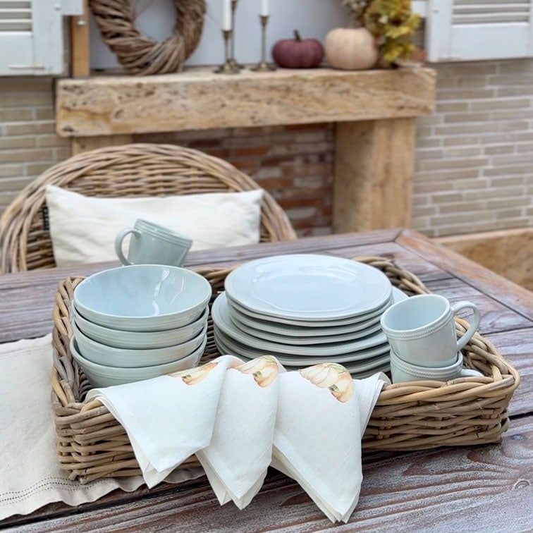 Set lichtgrijs keramisch servies met borden, kommen en mokken in een rieten mand op een houten tafel