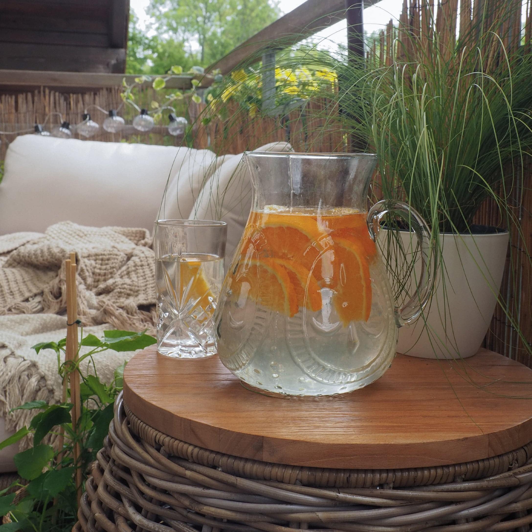 Glazen karaf met water en sinaasappelschijfjes op houten tafel op terras met planten