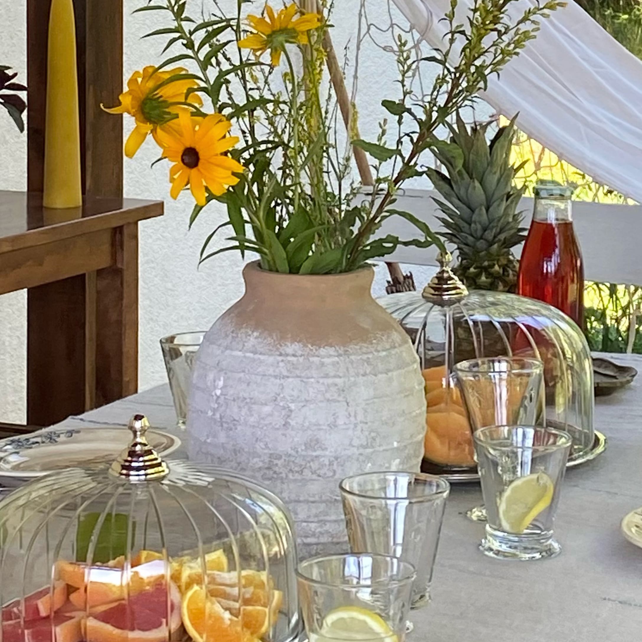 Beige-witte keramische vaas met gele bloemen op tafel met fruit en glazen.