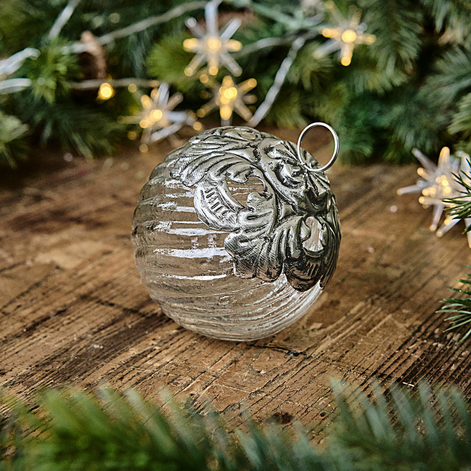 Kerstbal Zoriline Glazen kerstbal met zilveren metalen versiering op houten tafel met groene dennentakken
