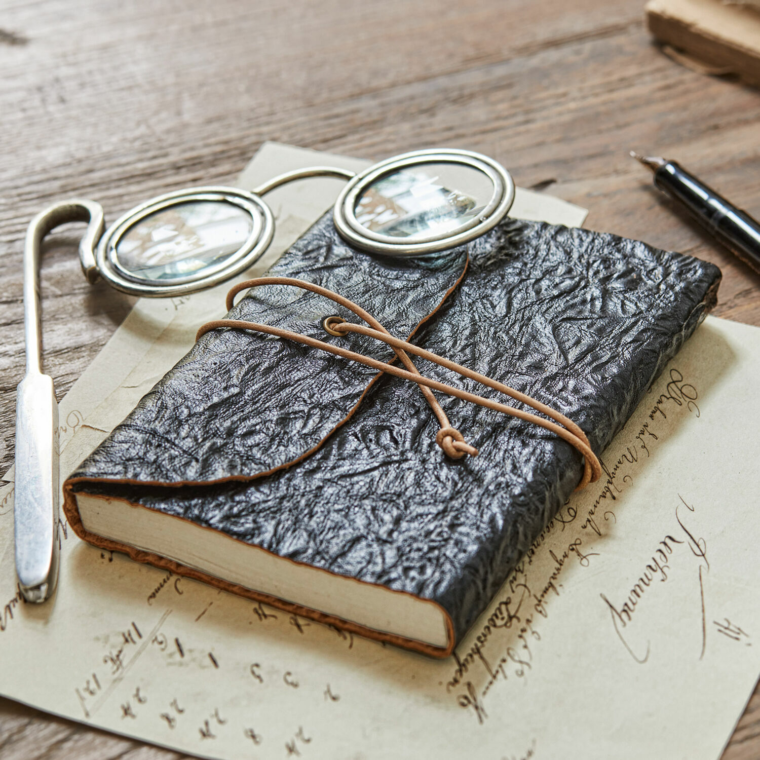 Notitieboekje Cosmes Zwart notitieboek van gestructureerd leer met koordsluiting op houten tafel met bril en pen