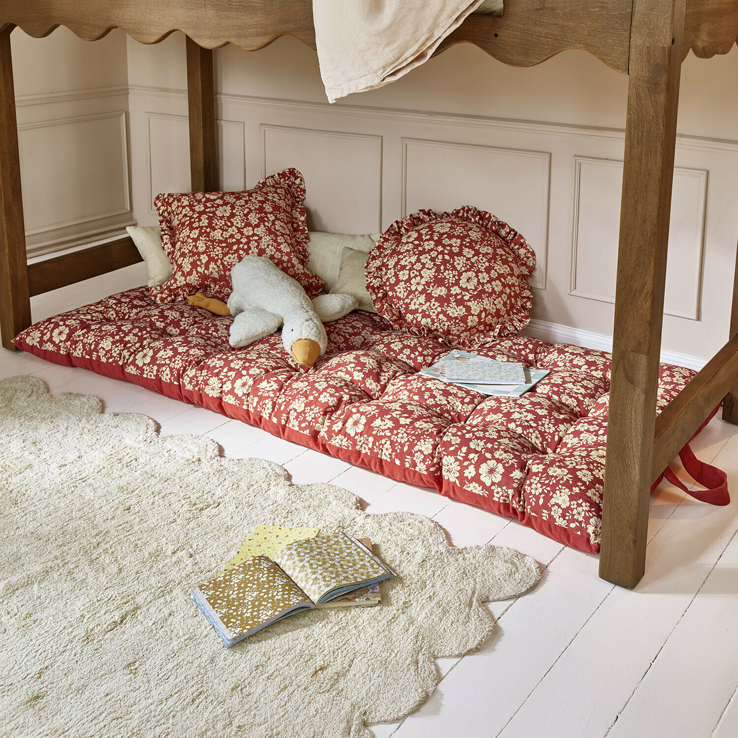 Rood matras met bloemenpatroon en kussens onder een houten tafel, knuffel en boeken op de vloer