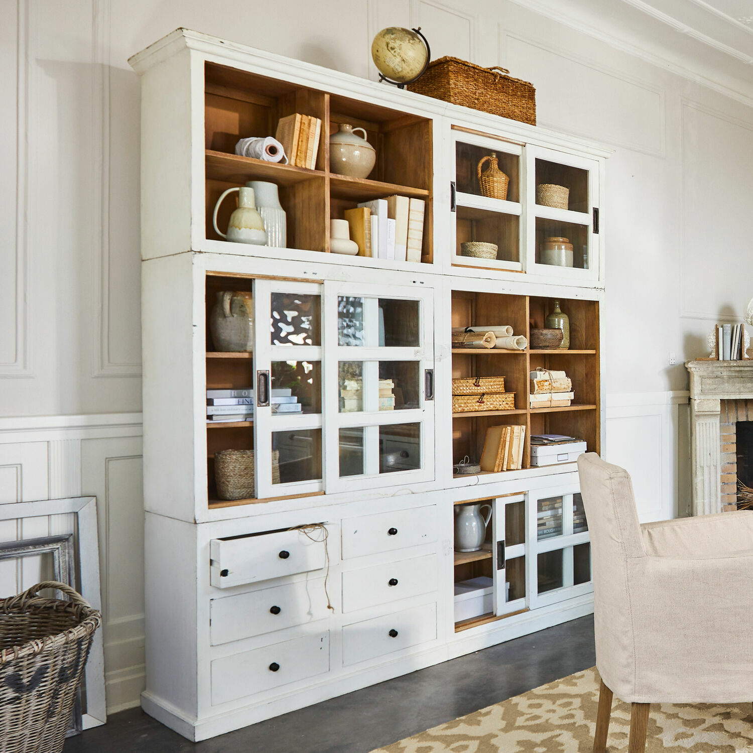 Kast Dubberly Witte houten kast met glazen deuren en lades in vintage stijl, versierd met boeken en manden