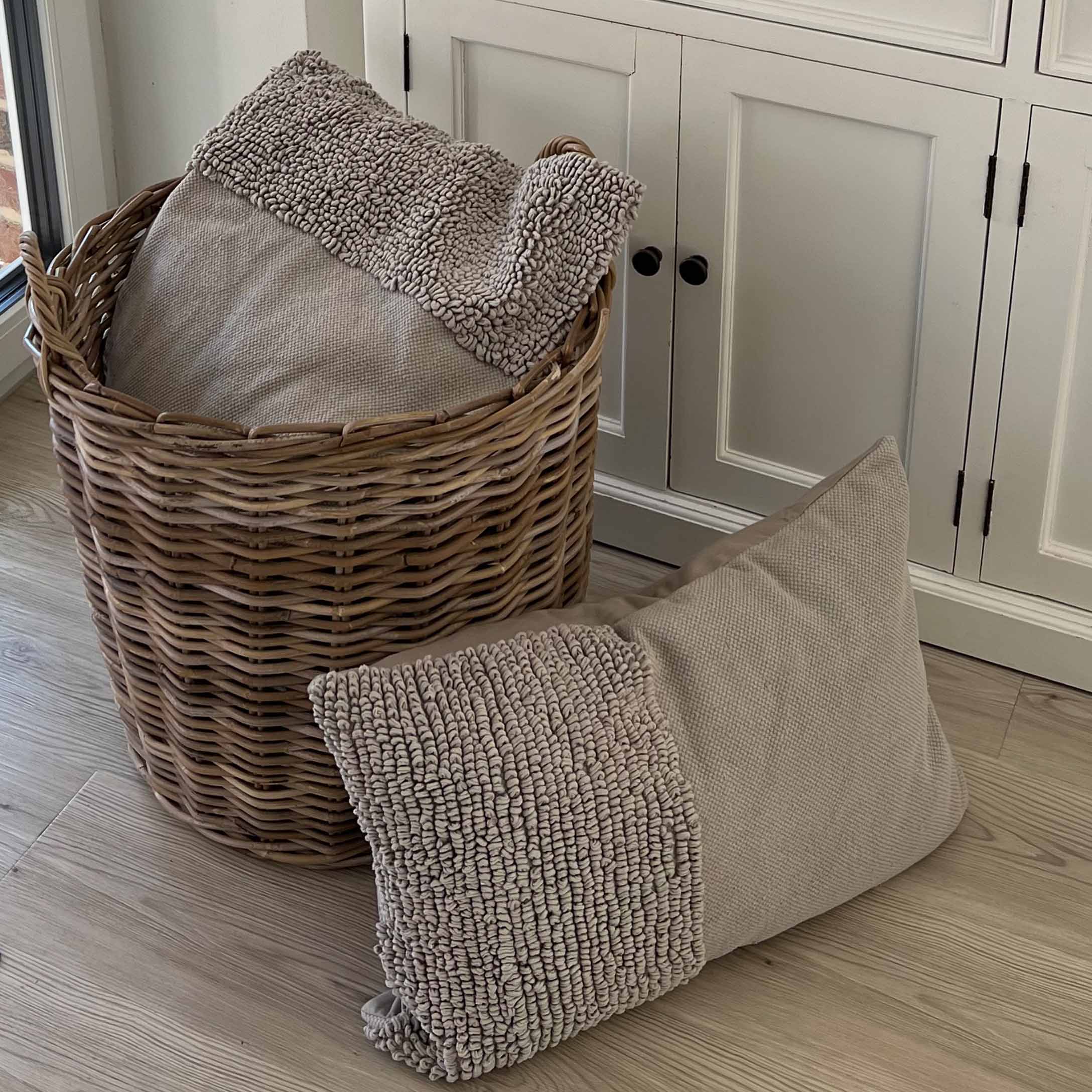 Beige kussen van zacht materiaal met textuur naast een rieten mand op een houten vloer