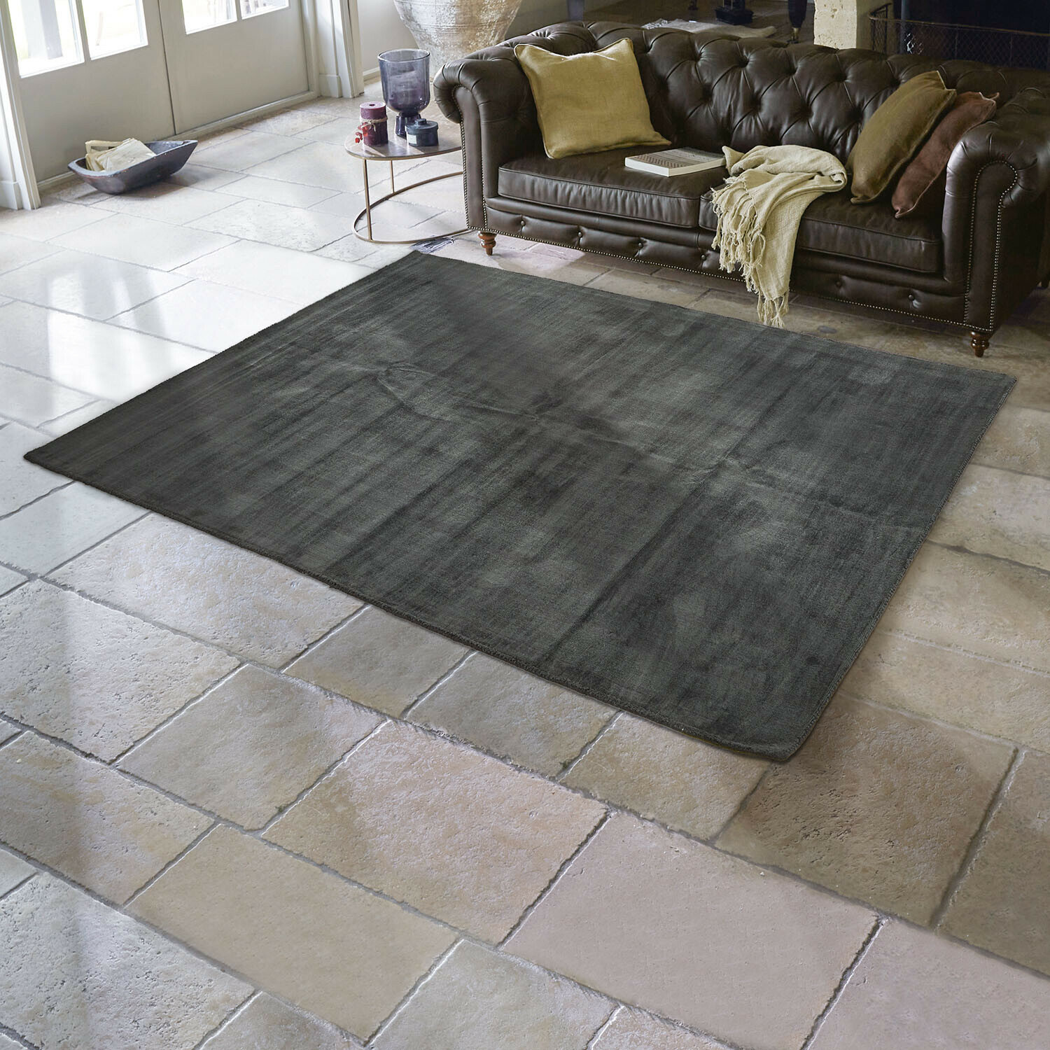Donkergrijs vloerkleed van zacht materiaal op stenen vloer in woonkamer met bruine leren bank