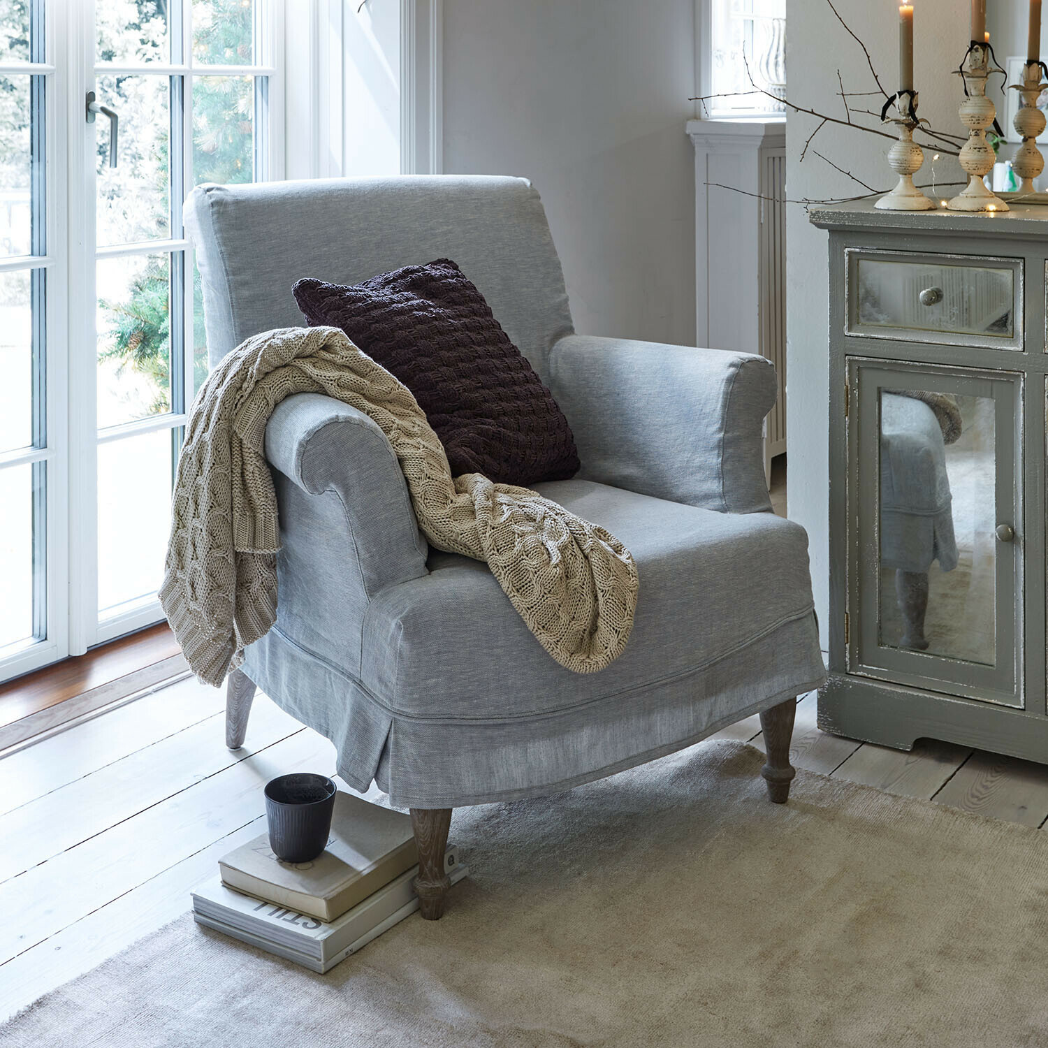 Grijze stoffen fauteuil met beige deken en donker kussen in een gezellige, lichte kamer