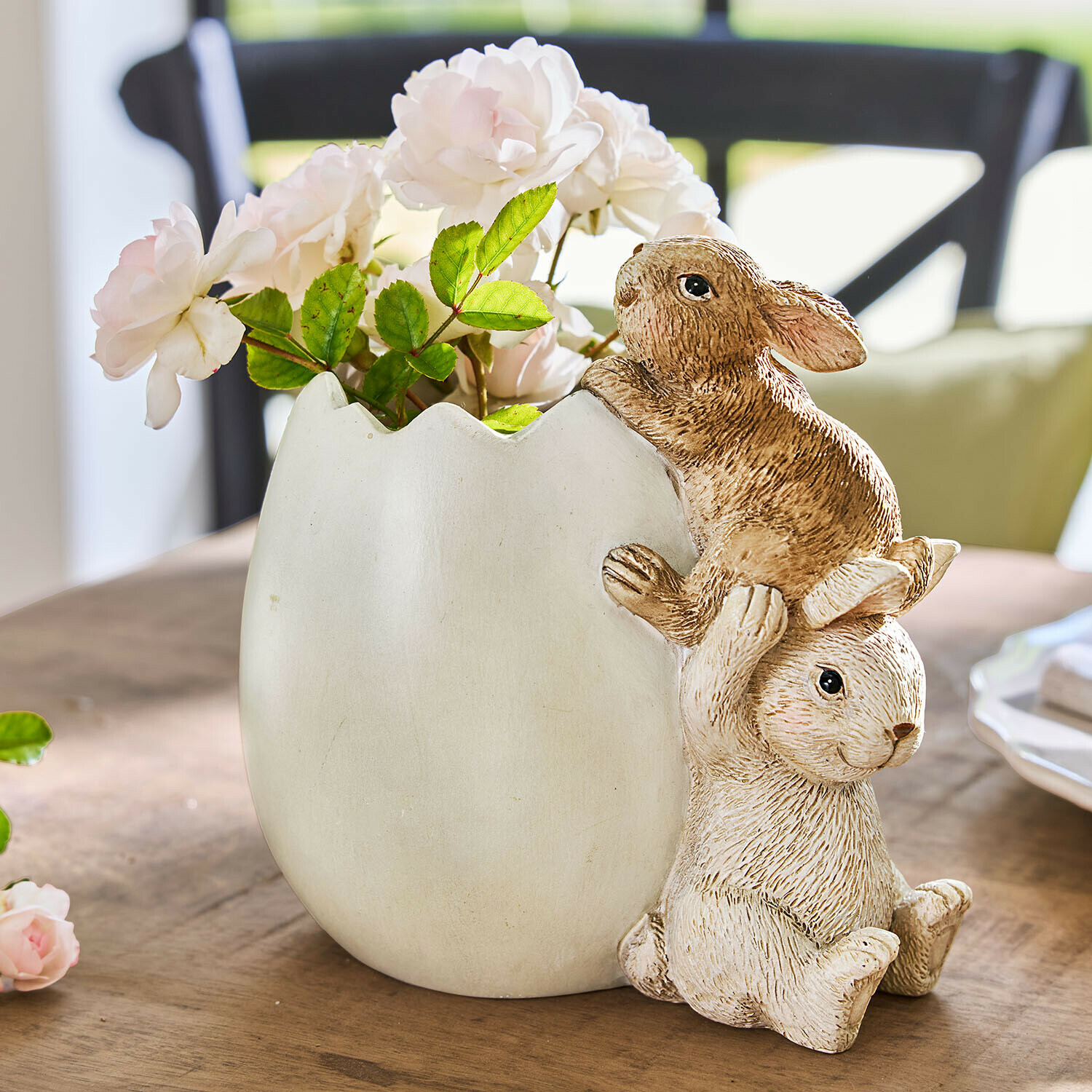 Witte keramische vaas in eierschaalvorm met twee konijnen en roze bloemen op houten tafel