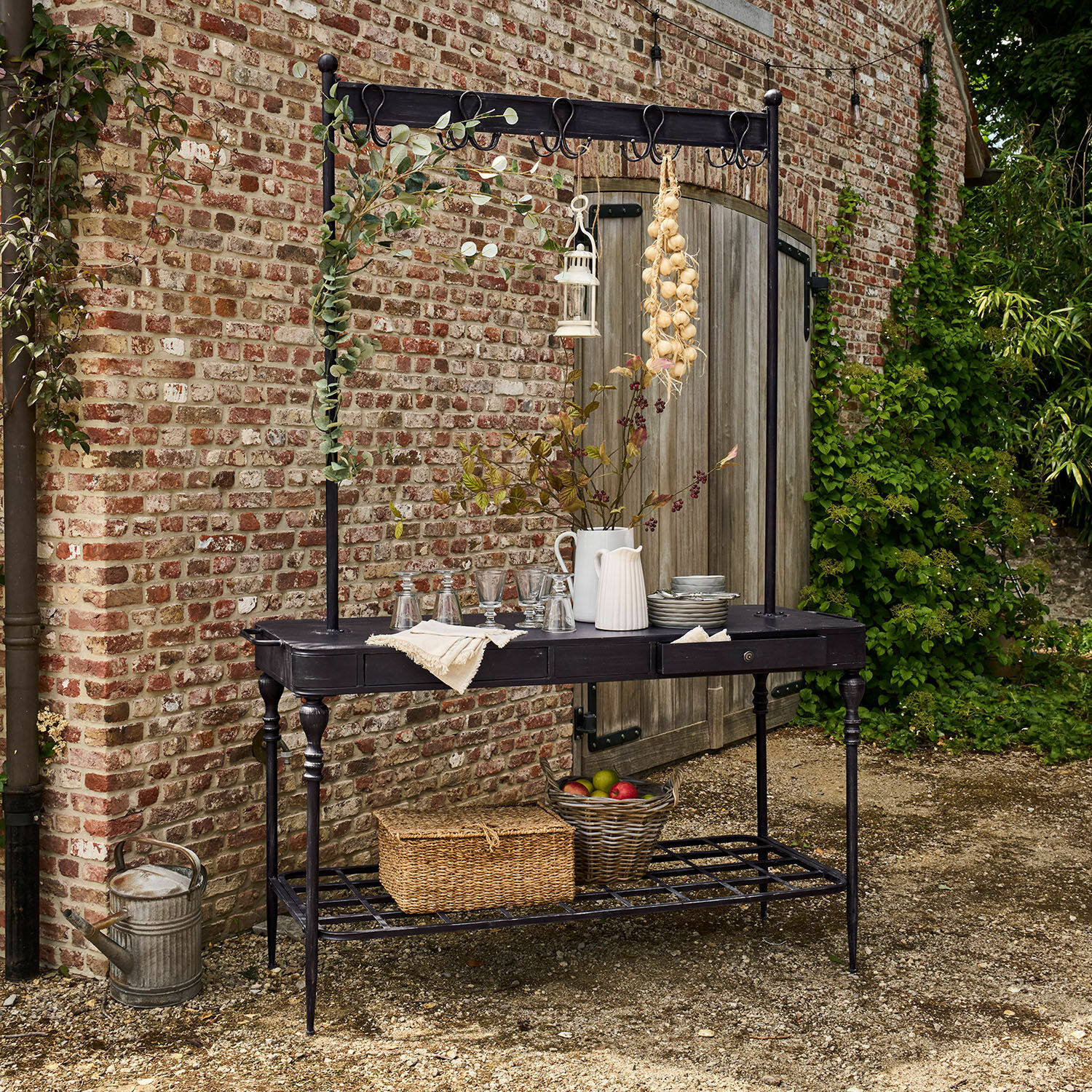 Rek Langmore Zwarte metalen tuintafel met haken en plank, versierd met kannen en fruitmand
