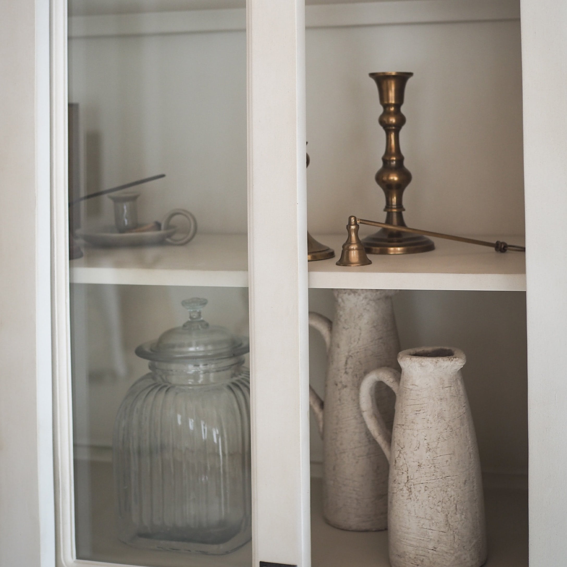 Glazen pot en witte keramische kannen in witte kast met messing kandelaar en belletje