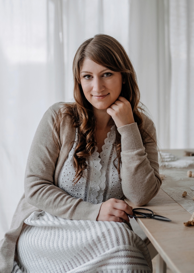 Vrouw in beige vest en witte kanten jurk zittend aan houten tafel met een schaar in de hand.