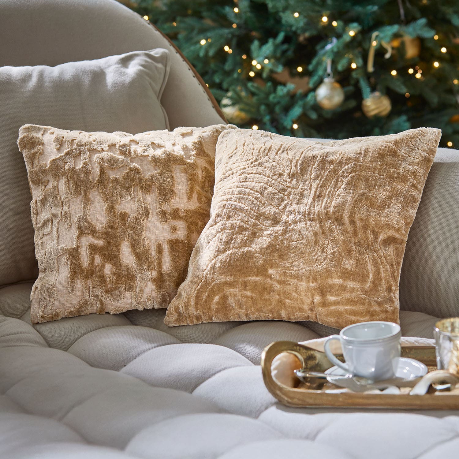 Twee beige decoratieve kussens van zacht materiaal op een lichte bank bij een versierde kerstboom
