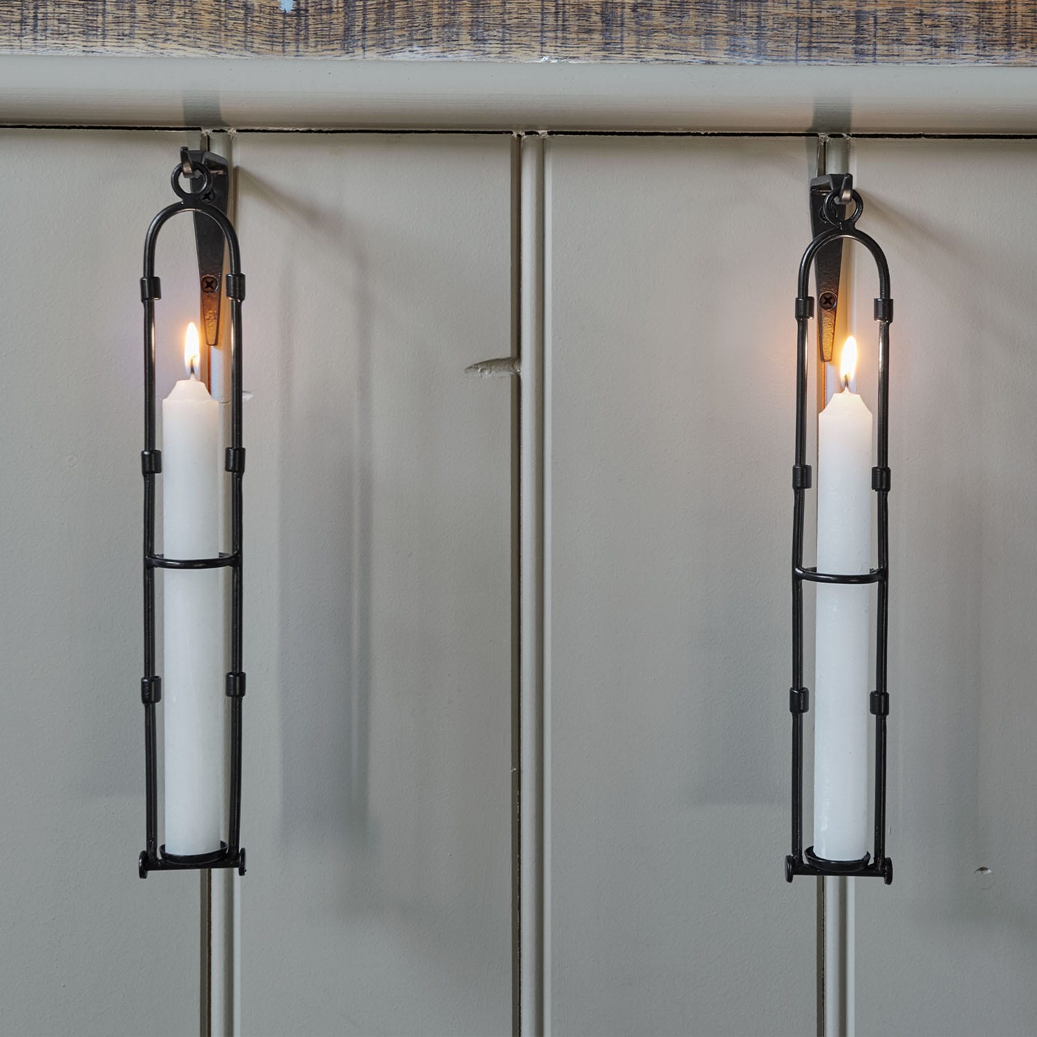 Twee zwarte metalen wandkandelaars met brandende witte kaarsen aan een lichtgrijze muur