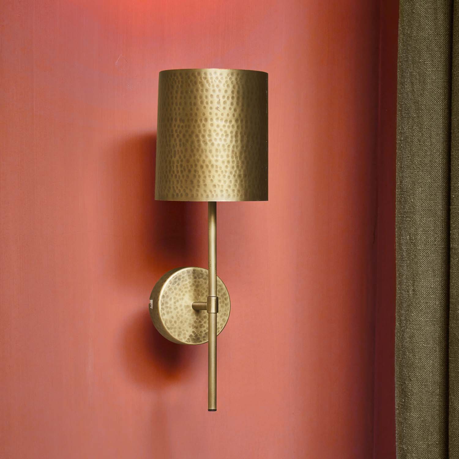 Messing wandlamp met hamerslagpatroon en cilindrische kap aan rode muur, decoratieve verlichting