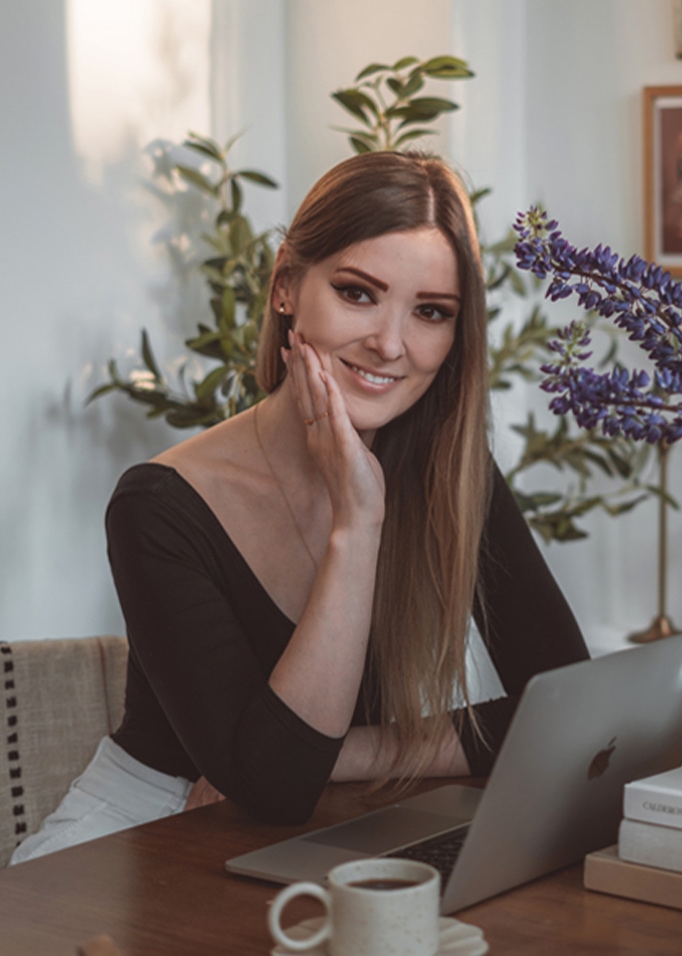Vrouw zittend aan houten tafel met laptop, koffiekopje en bloemen op de achtergrond
