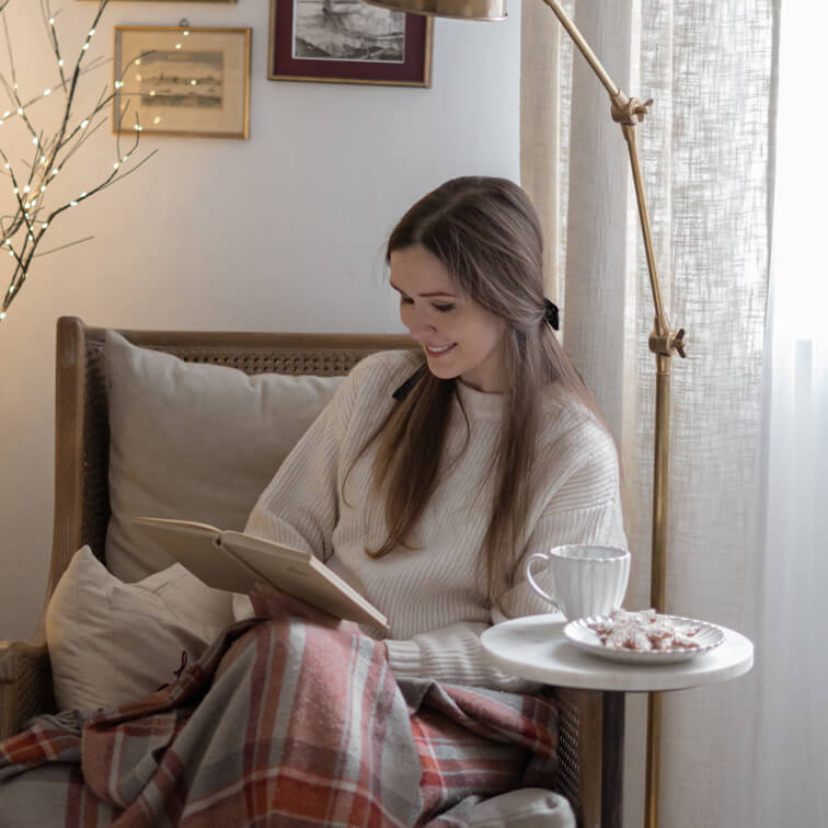 Vrouw leest boek in rotan stoel met crèmekleurige trui en warme geruite deken