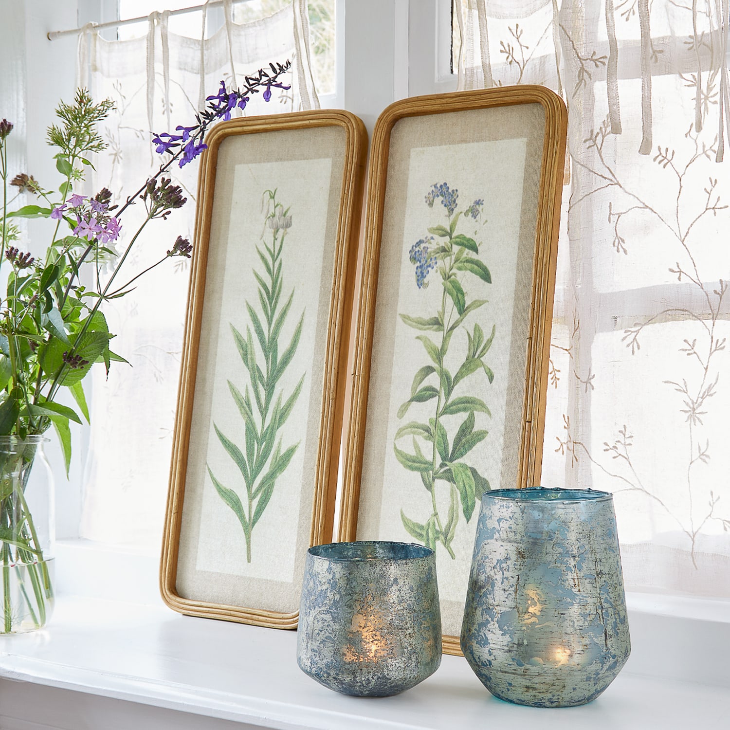 Twee lijsten met botanische illustraties en blauwe glazen windlichten op vensterbank met gordijnen