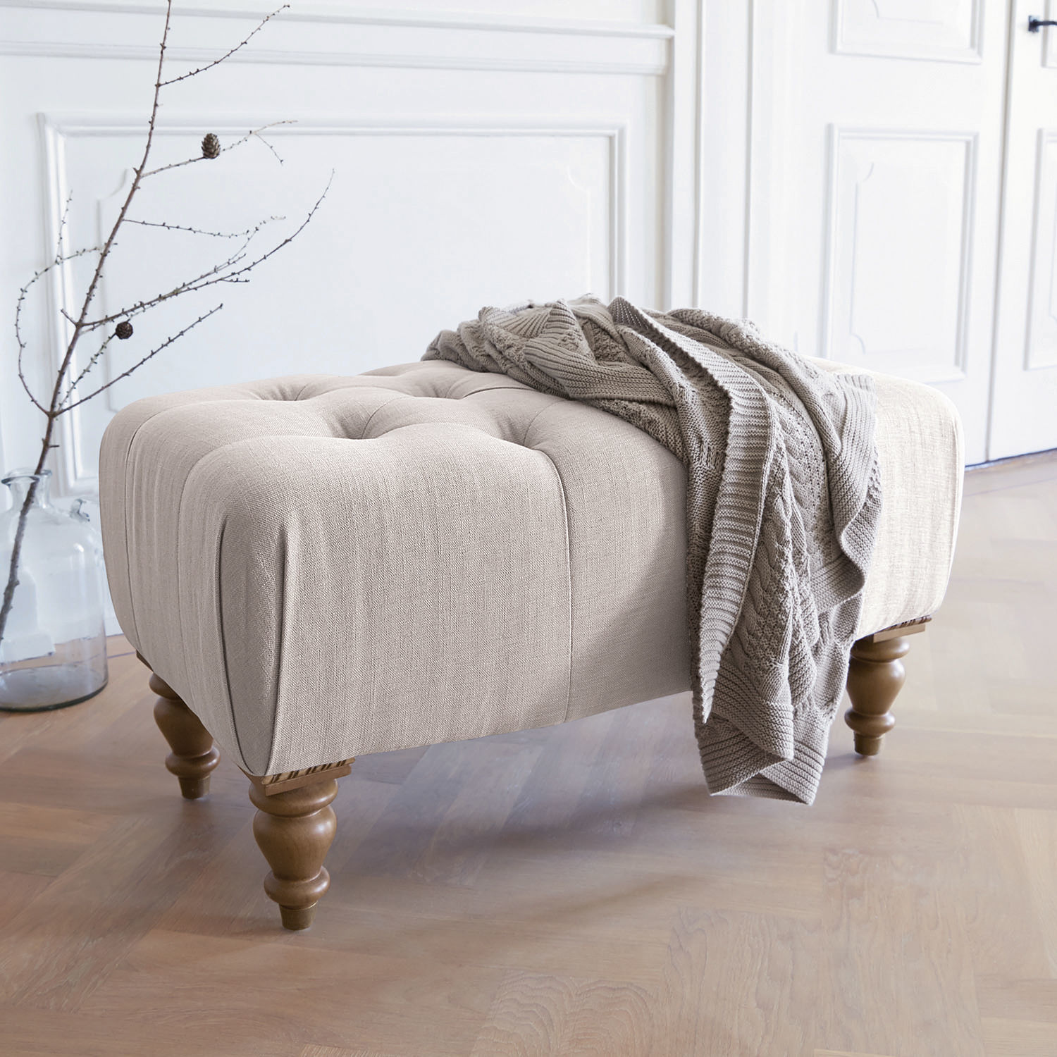 Hocker Durance Beige gestoffeerde poef met houten poten en grijze gebreide deken in lichte kamer