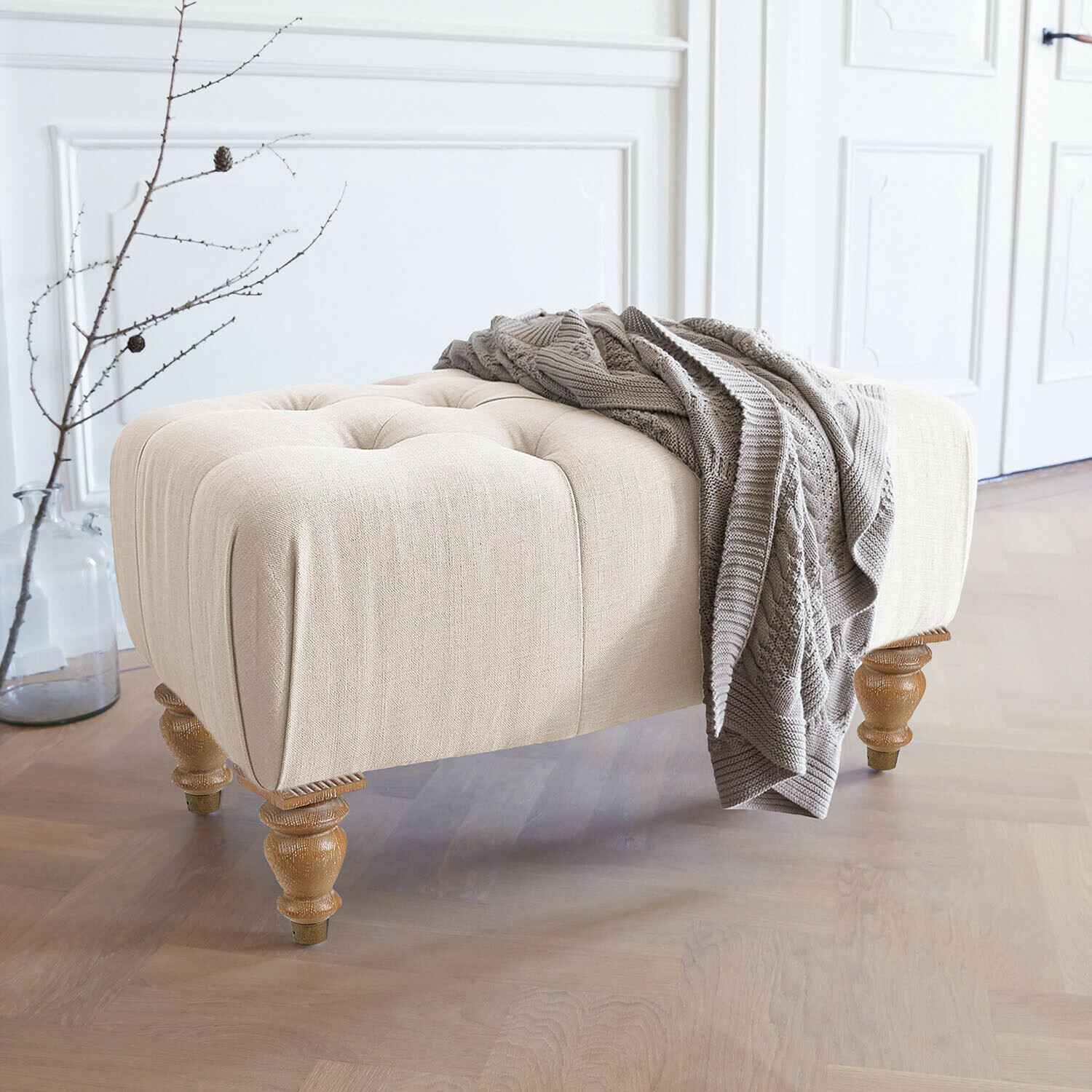 Hocker Durance Beige gestoffeerde poef met houten poten en grijze gebreide deken in lichte kamer
