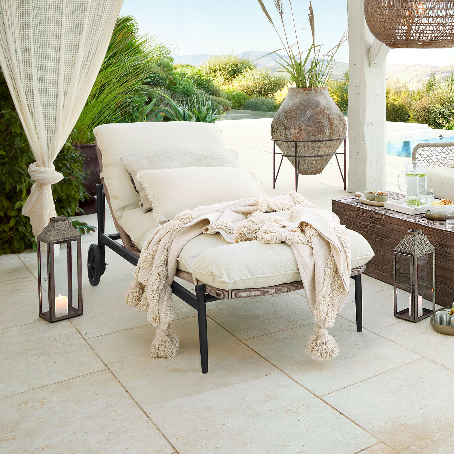 Tuinligstoel met crèmekleurige kussens en zachte deken op terras met uitzicht op zwembad en groen