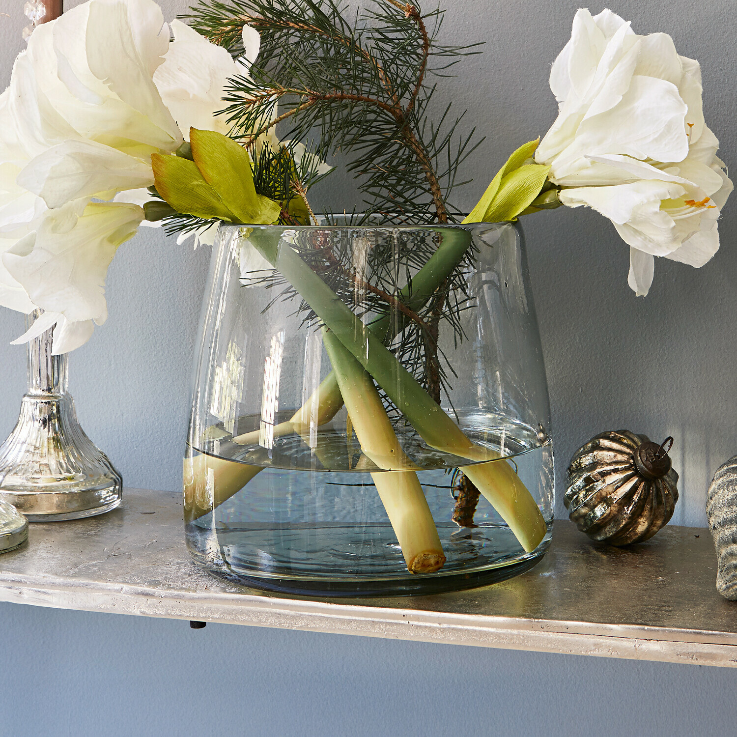 Vaas Inga Transparante glazen vaas met witte bloemen en groene takken op een decoratieve plank