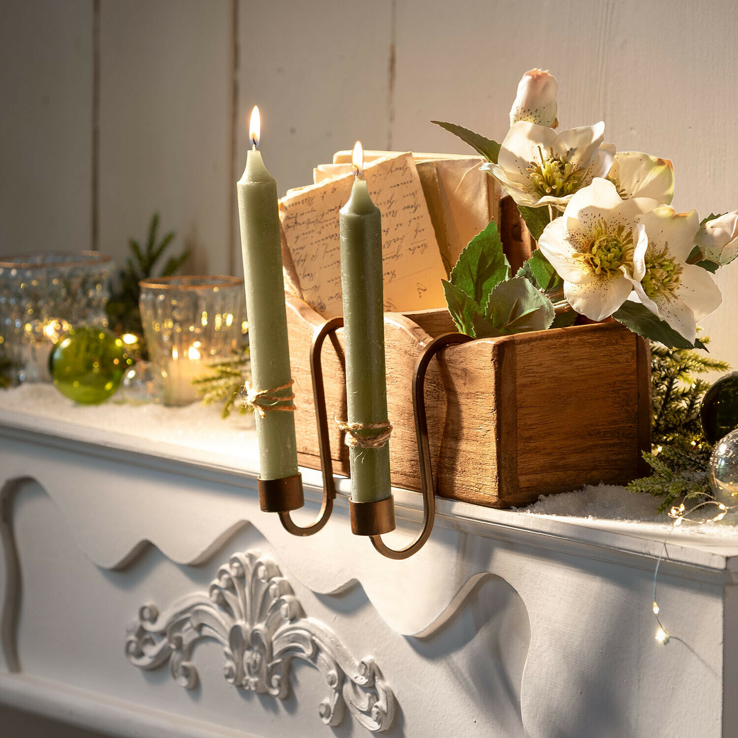 Dubbele messing kandelaar met groene kaarsen op een witte schoorsteenmantel met bloemen en houten doos