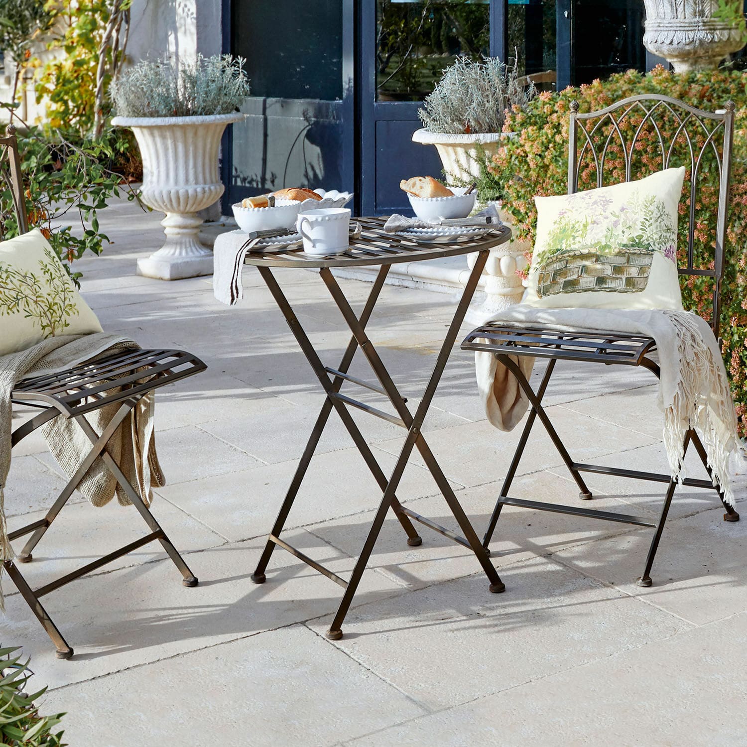 Tafel Brignoles Bruine metalen inklapbare tafel en stoelen op terras met decoratieve kussens en wit servies