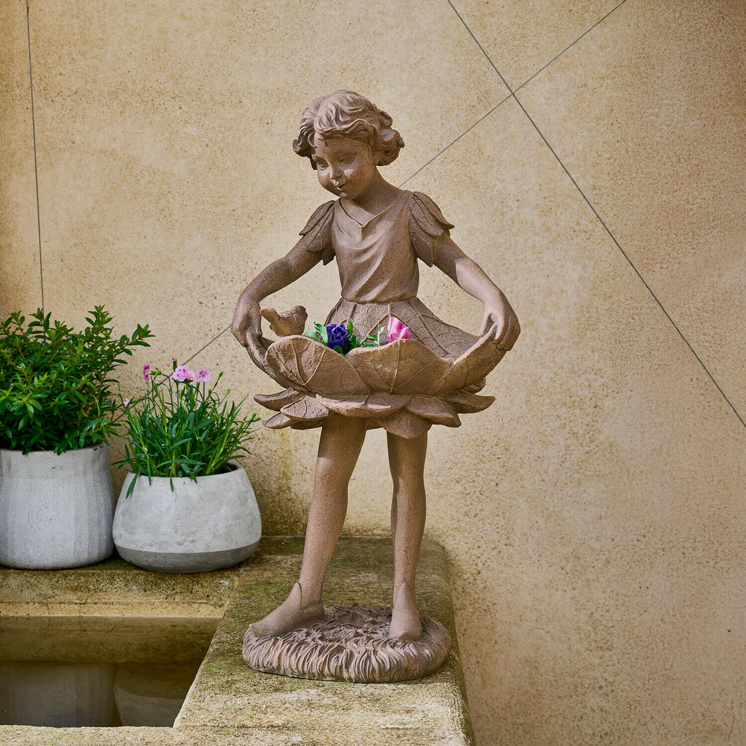 Beeld van een meisje van bruin steen met een bloemenschaal en kleurrijke bloemen in de tuin