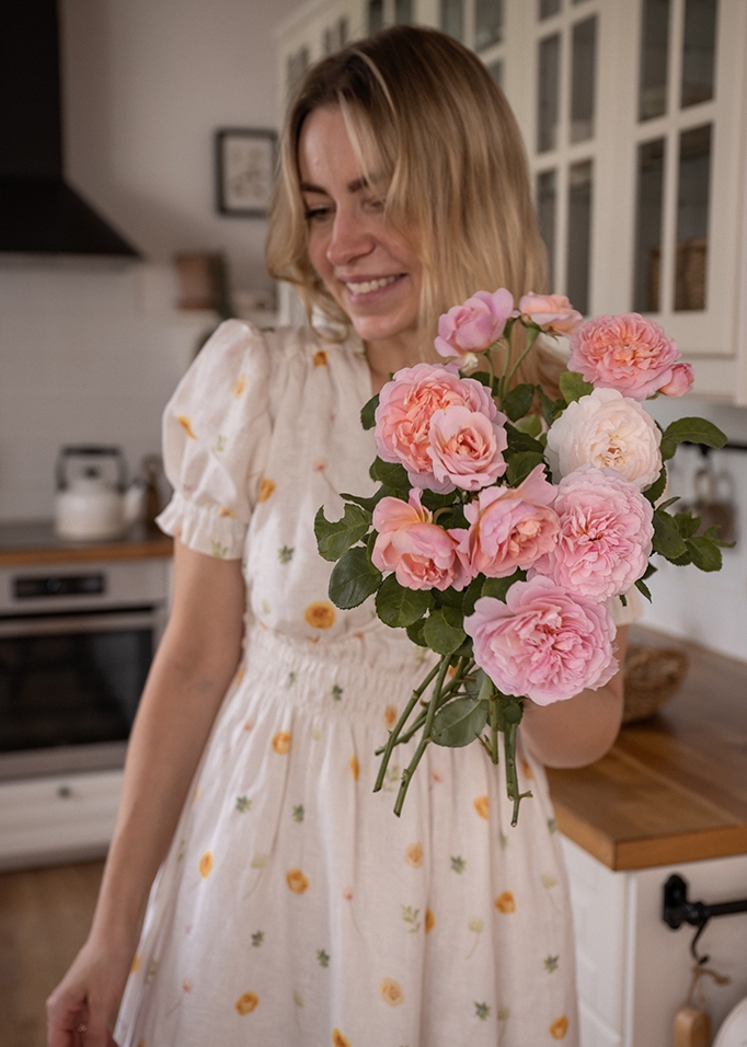 Vrouw in witte jurk met bloemenpatroon houdt een bos roze rozen vast in de keuken