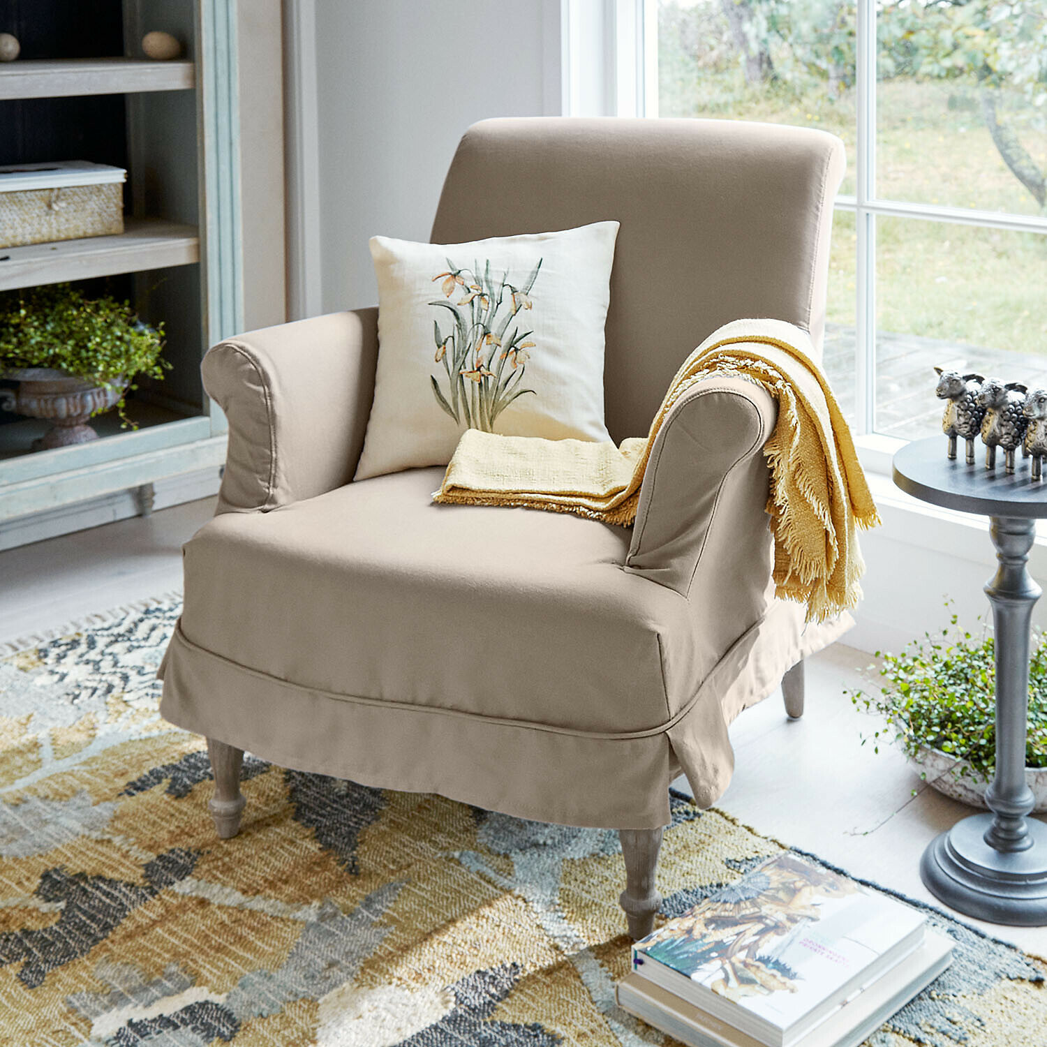 Beige stoffen fauteuil met bloemkussen en gele deken in lichte woonkamer met kleurrijk vloerkleed