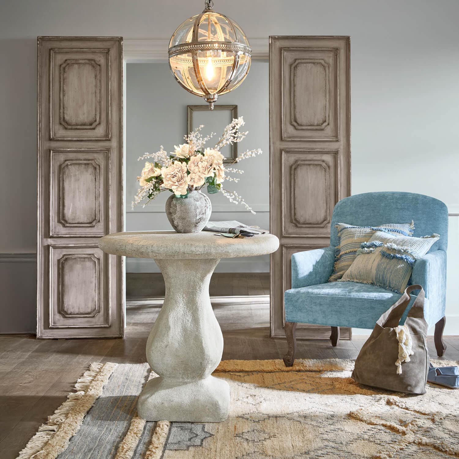 Ronde grijze betonnen tafel met bloemenvaas, blauwe fauteuil en bruine tas in lichte kamer