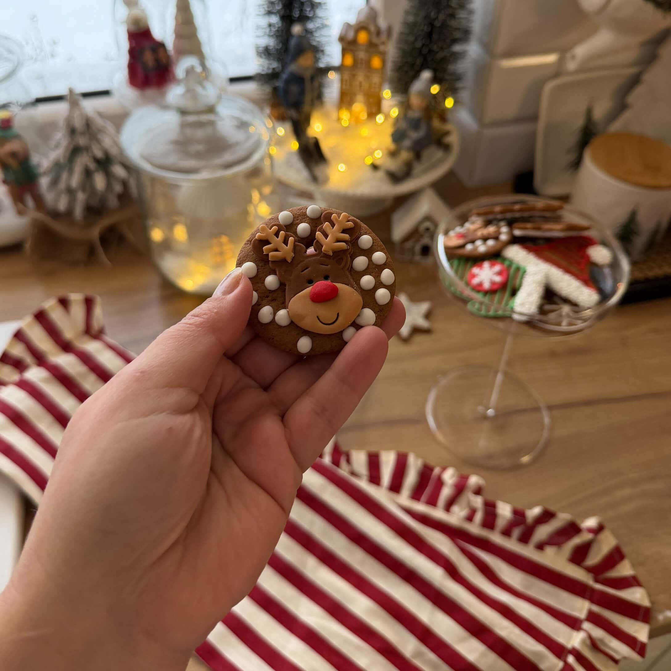 Hand houdt peperkoek met rendier en witte stippen, kerstdecoraties en koekjes op de achtergrond